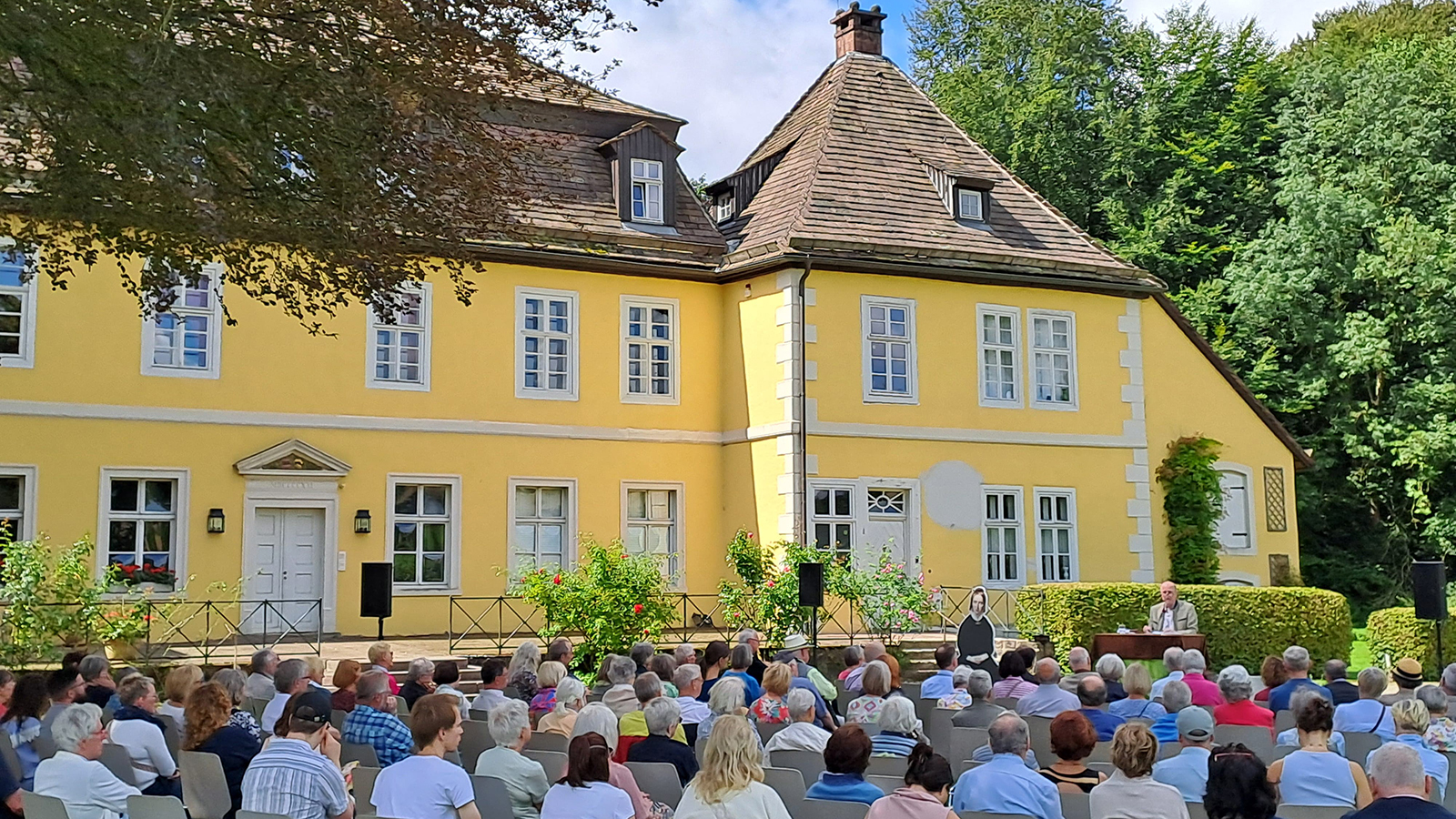 Die strahlende Sonne verstärkte den schönen, gelben Farbton des spätbarocken Hauses Bökerhof, der Himmel im satten Blau, der Rasen im frischen Grün, ein laues Lüftchen wehte: in diesem idyllischen Ambiente im Schatten einer riesigen Blutbuche referierte Wilderich Freiherr von Droste zu Hülshoff sehr kenntnisreich über seine berühmte Ur-Urgroßtante Annette